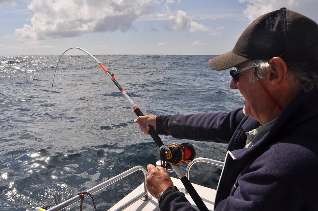 En fisker på en båt som aktivt kjemper mot en fisk med en bøyd fiskestang i åpent hav. Personen har på seg passende fiskeantrekk inkludert caps og solbriller. Bildet fanger spenningen ved sjøfiske med en vakker horisont og havutsikt. Den bøyde stangen indikerer at fisken gjør motstand.